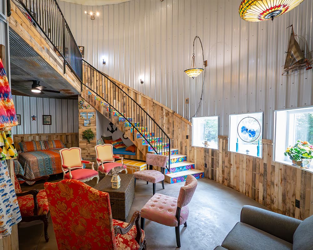 Interior of a colorful, rustic living space featuring a spiral staircase, cozy seating, and large windows.