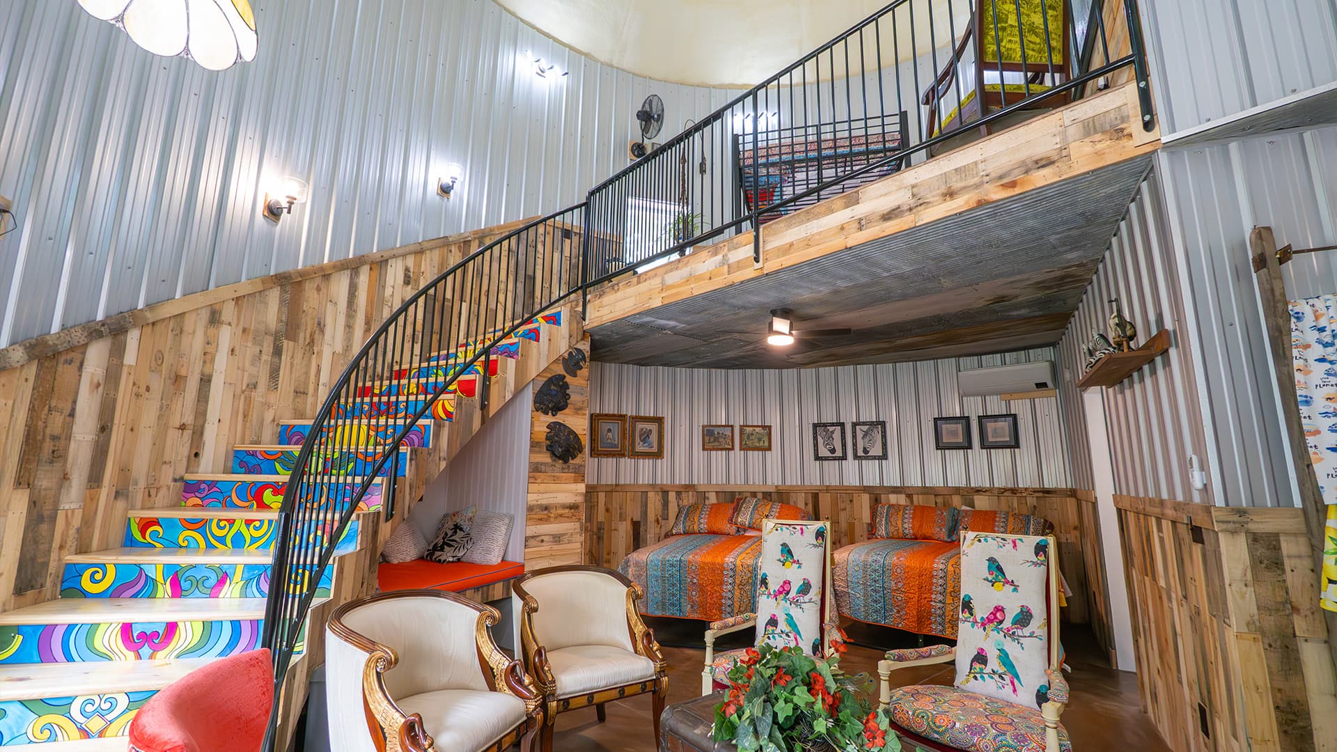 Colorful, rustic interior of a circular room featuring a spiral staircase, cozy seating, and patterned bedding.
