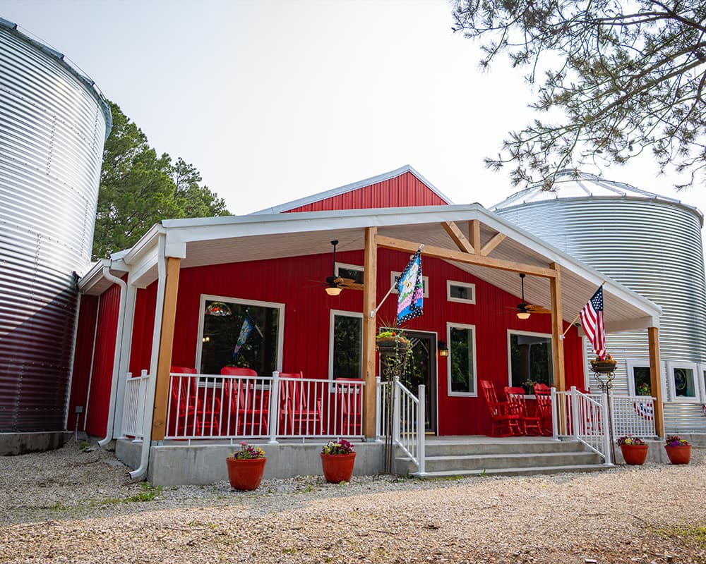 Red farmhouse with a porch, surrounded by silos and potted plants.